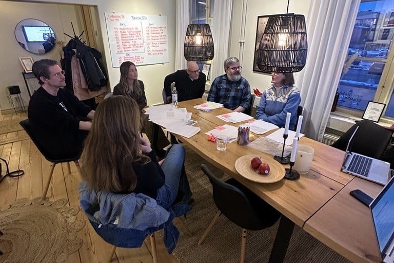 Sex personer män och kvinnor sitter runt ett bord fullt med papper.